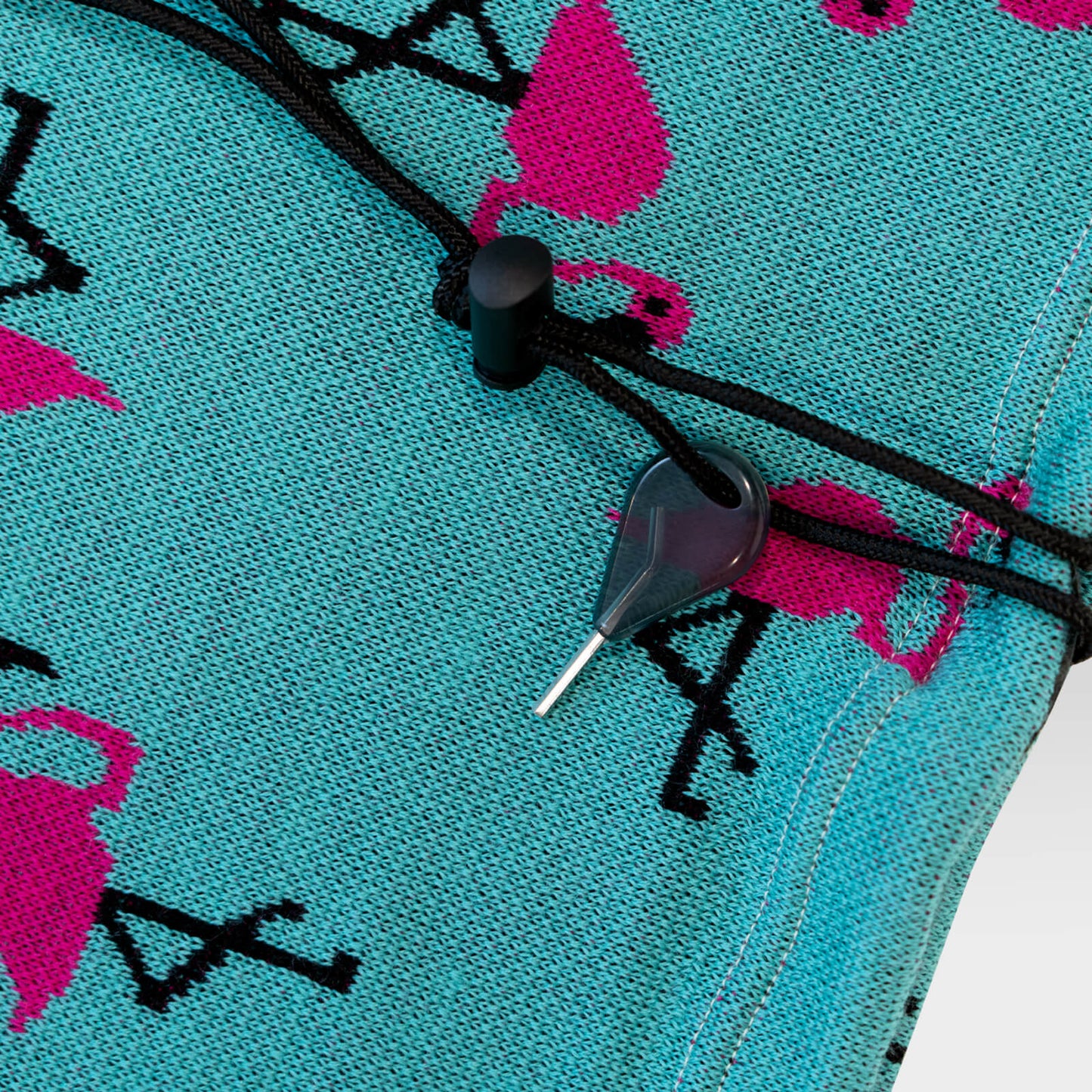 Close-up of a turquoise knit fabric with a pink and black flamingo pattern, featuring a black cord closure and a small black plastic tool.